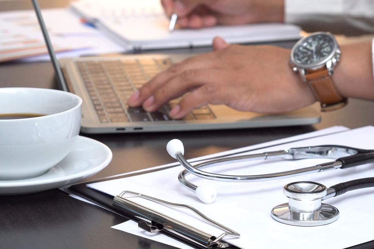 Medical professional typing on a laptop, with a coffee cup, stethoscope and clipboard on the table